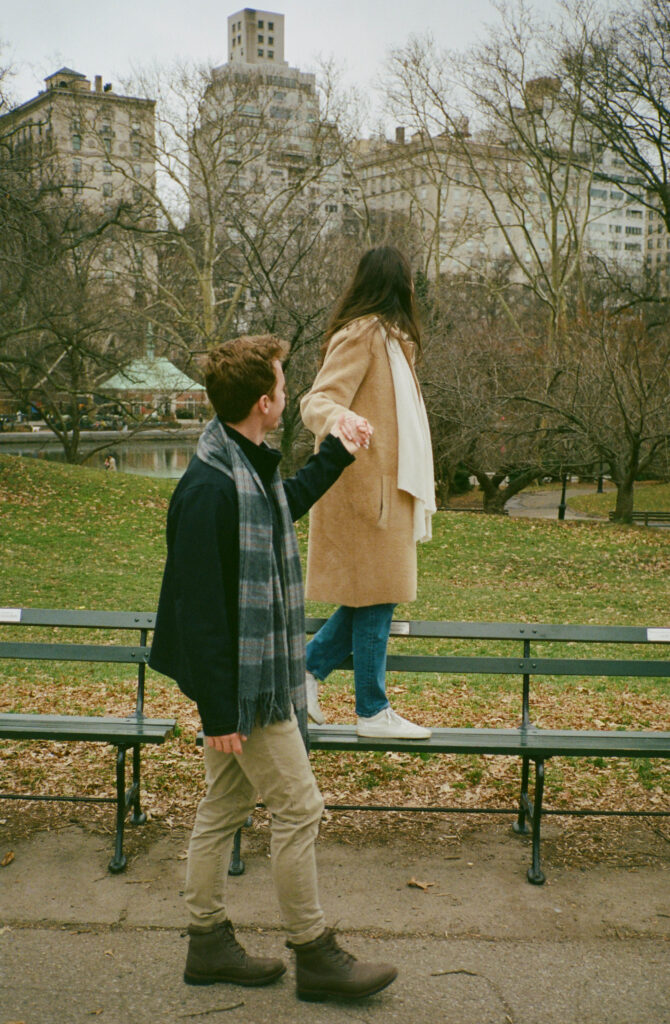 A couple stands at a bench in Central Park. Discover candid film photos from an engagement session in New York City captured on 35mm film.  