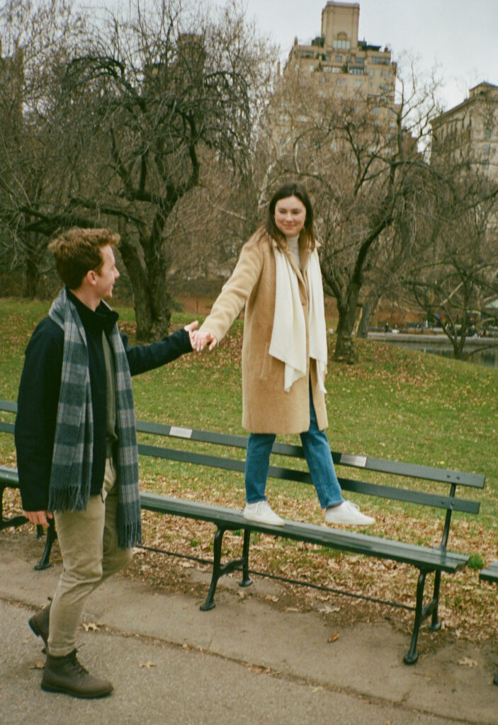 A couple stands at a bench in Central Park. Discover romantic film photos from an engagement session in New York City captured on 35mm film.  