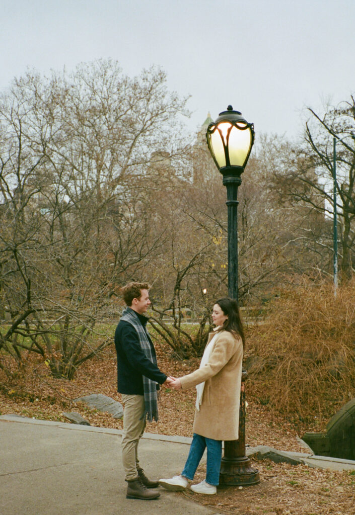 A couple stands together at a light post in Central Park. Discover documentary film photos from an engagement session in New York City captured on 35mm film.  