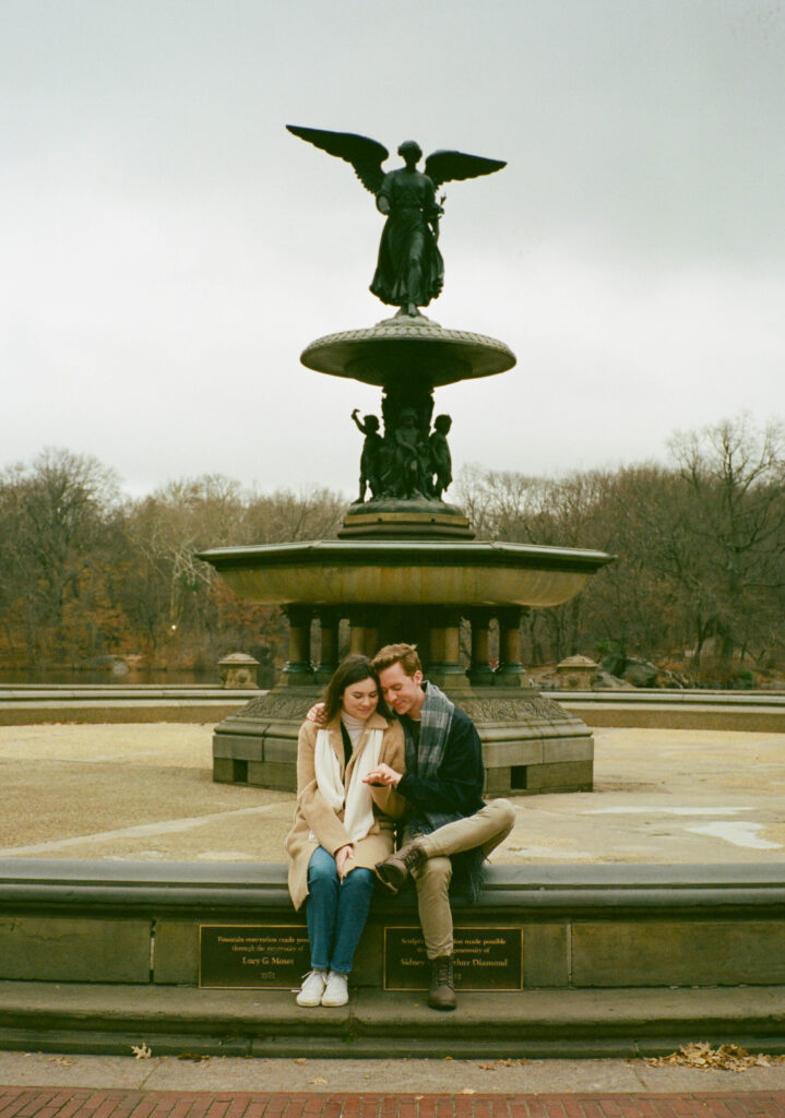 A couple sits together at Bethesda Fountain in Central Park. Discover intimate film photos from an engagement session in New York City captured on 35mm film.  