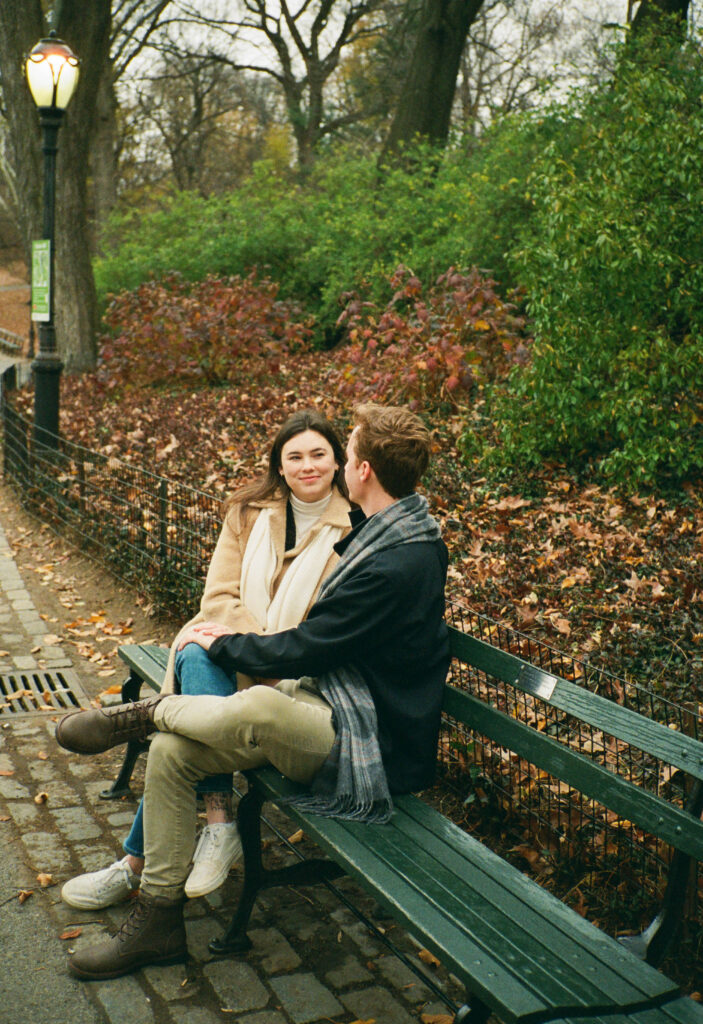 A couple sits together at a bench in Central Park. Discover romantic film photos from an engagement session in New York City captured on 35mm film.  
