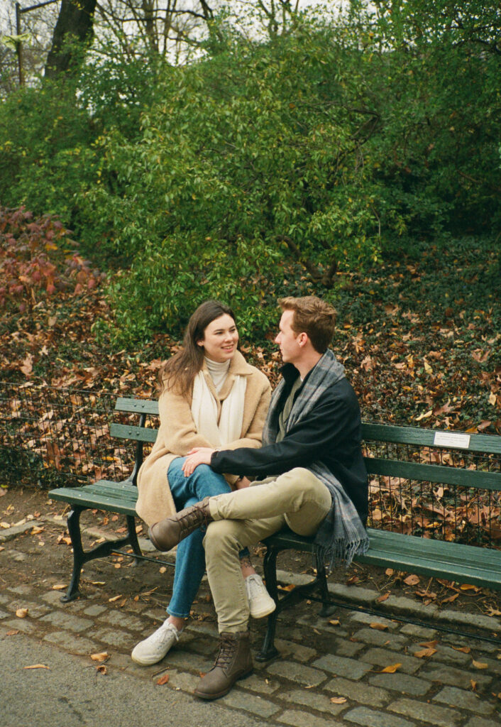 A couple sits together at a bench in Central Park. Discover romantic photos from an engagement session in New York City captured on 35mm film.  