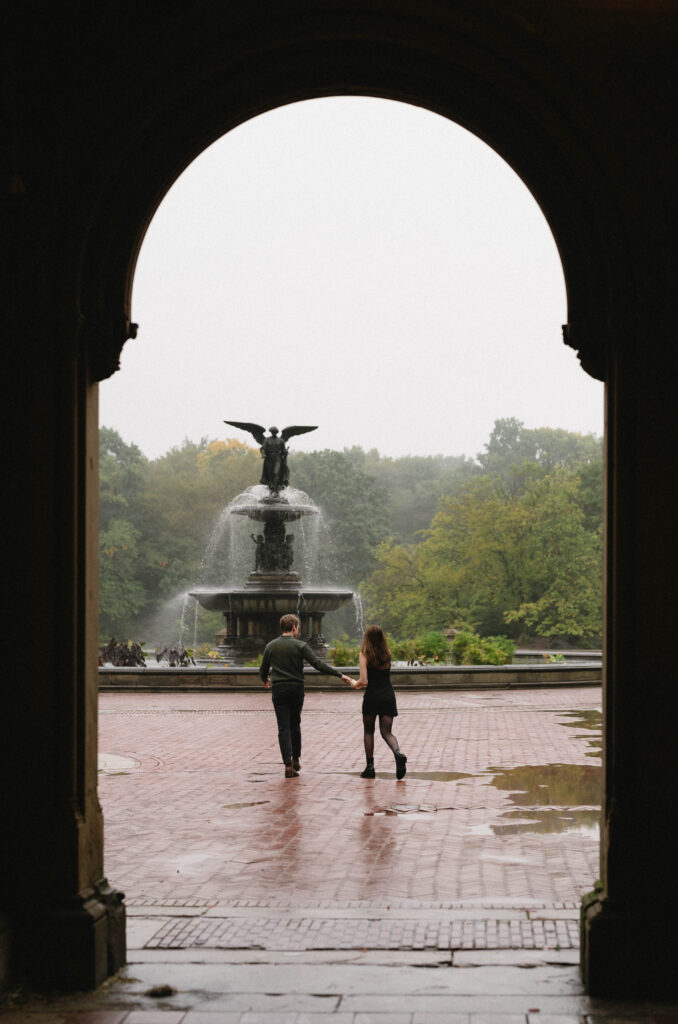 A documentary photo of a couple running through the rain toward Bethesda Fountain in central park. A candid moment captured for their intimate engagement session in New York City. 