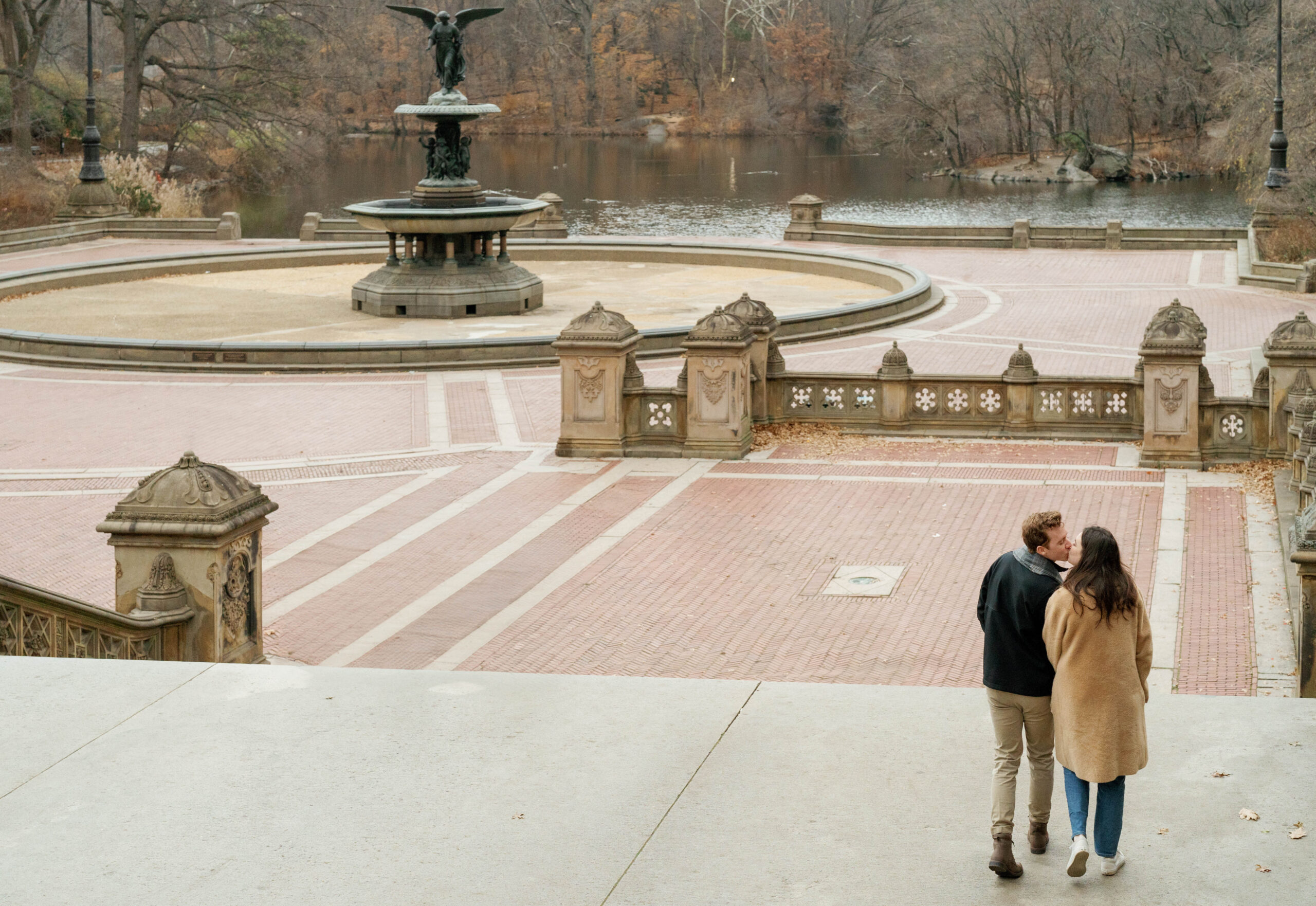 A couple kisses while walking down the stairs of Bethesda Terrace towards Bethesda fountain at their engagement session in New York City.